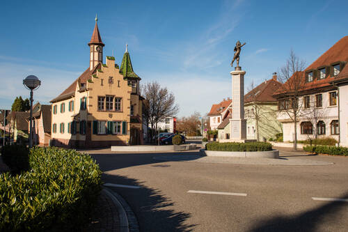 Ein idyllischer Platz mit einem historischen Rathausgebäude mit spitzen Türmen und einer Statue, die auf einer Säule steht, ist von Büschen und weiteren Gebäuden umgeben.