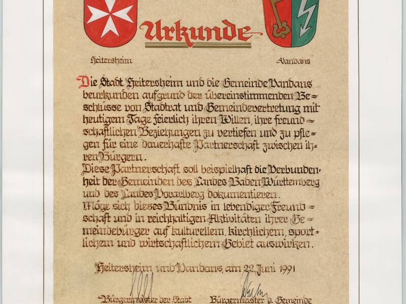 Das Bild zeigt eine Urkunde, die die Partnerschaft zwischen der Stadt Heitersheim und der Gemeinde Vandans feiert, versehen mit zwei farbigen Wappen und handschriftlichen Unterschriften.