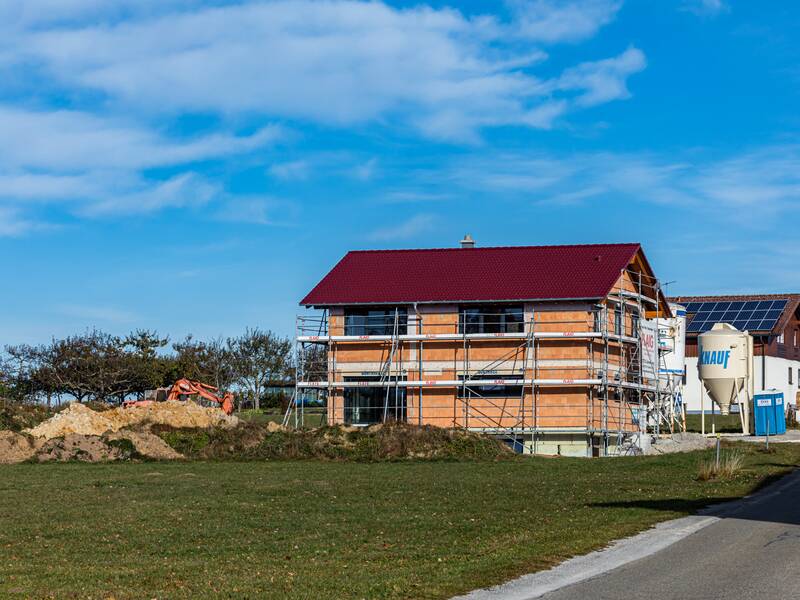 Neu gebautes Haus, das noch eingerüstet ist. Das Haus steht am Ende eines Wohngebiets auf einer grünen Wiese. Der Himmel ist blau.